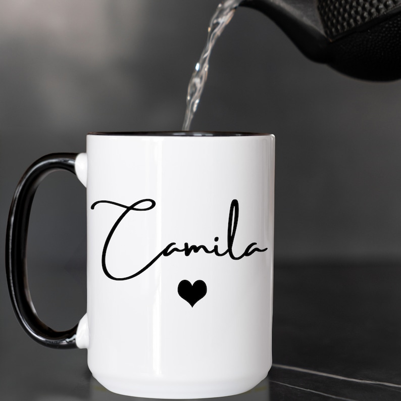 May include: A white ceramic mug with a black handle and interior rim. The front of the mug features the name Camila written in a black cursive script font, positioned above a small, solid black heart icon. Liquid is being poured into the mug.