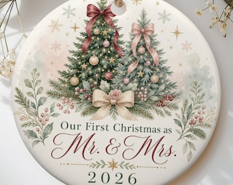 May include: A round white ceramic Christmas ornament featuring two illustrated pine trees decorated with ornaments and bows. The text reads Our First Christmas as Mr. & Mrs. 2026. The design includes watercolour greenery, red berries, and gold snowflakes.