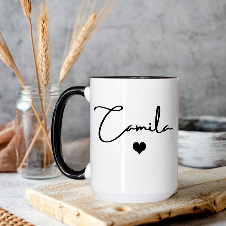 May include: A white ceramic mug with a glossy finish, featuring a black handle and a black interior rim. The front of the mug displays the name Camila written in a black cursive script font, positioned directly above a small, solid black heart icon.