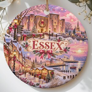 May include: A round ceramic ornament featuring a winter scene of a castle and a coastal town with the text ESSEX in the center. The image includes snow-covered buildings, holiday lights, a ferris wheel, and a signpost with the words Colchester, Castle, History, and Essex.