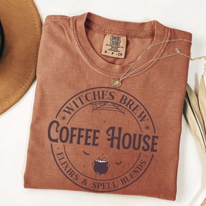 以下が含まれることがあります： くすんだ茶色のTシャツに、円形のグラフィックがプリントされています。グラフィックには「Witches Brew Coffee House Elixirs & Spell Blends」という文字と、釜、コウモリ、クモの巣が描かれています。