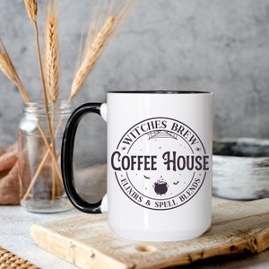 May include: White ceramic mug with a black handle and black rim. The mug has a black and white graphic of a cauldron, bats, and the text "Witches Brew Coffee House - Elixirs & Spell Blends".