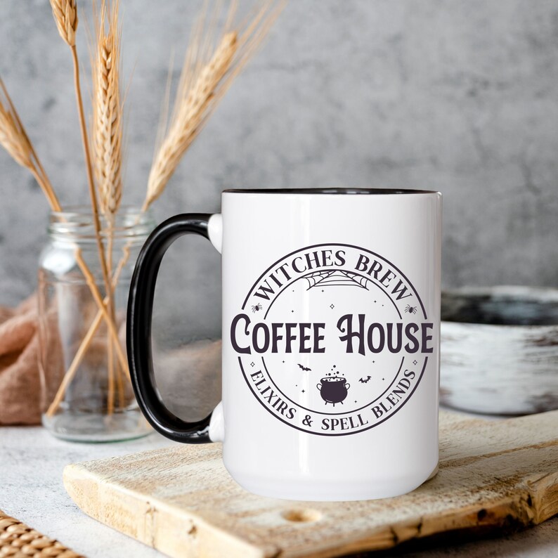 May include: White ceramic mug with a black handle and black rim. The mug has a black and white graphic of a cauldron, bats, and the text "Witches Brew Coffee House - Elixirs & Spell Blends".