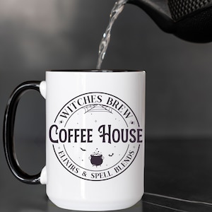 May include: White coffee mug with a black handle and black rim. The mug has a black and white graphic that says "Witches Brew Coffee House Elixirs & Spell Blends" with a cauldron and bats.