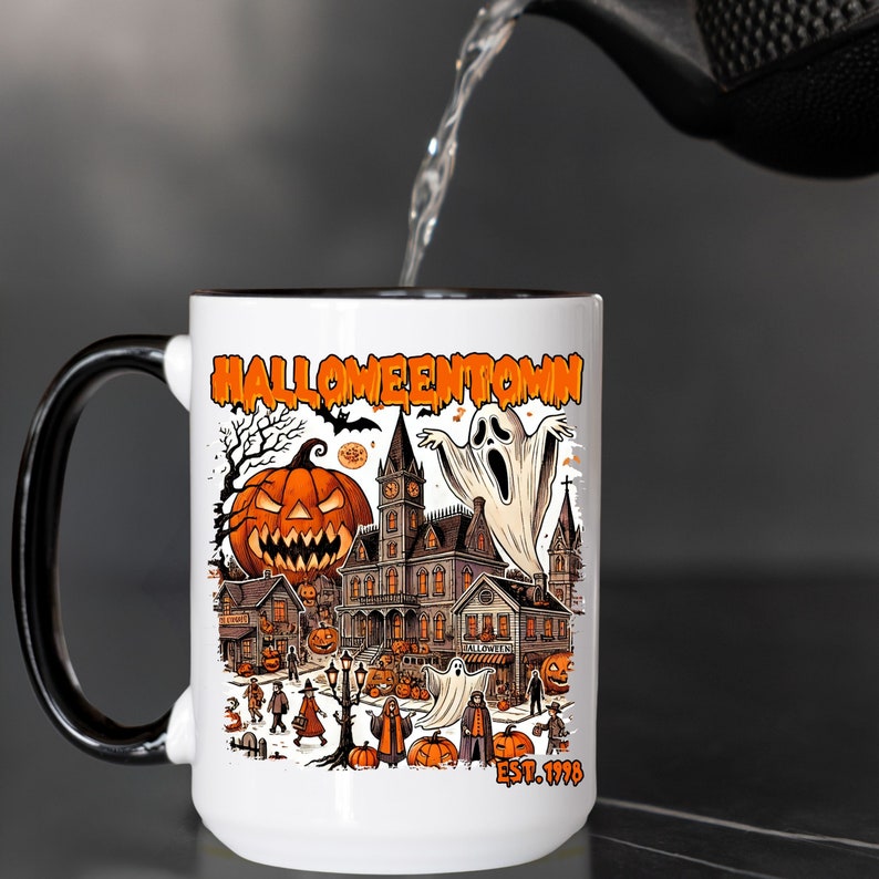 May include: White ceramic coffee mug with a black handle. The mug has a black rim and features a colorful illustration of a Halloween town with a ghost, a pumpkin, and the text "Halloweentown Est. 1998".