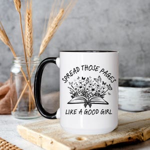 Puede incluir: Taza de cer&aacute;mica blanca con asa y borde negros. La taza presenta el texto "Spread Those Pages Like a Good Girl" con un dise&ntilde;o floral y de mariposas. La taza est&aacute; sobre una superficie de madera.