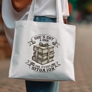 Może przedstawiać: Biała torba typu tote z grafiką stosu książek i tekstem "She's got a book for every situation".