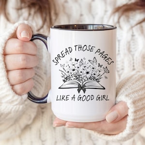 May include: White ceramic mug with a black interior and handle. The mug features the text "SPREAD THOSE PAGES LIKE A GOOD GIRL" with a graphic of an open book, flowers, and butterflies.
