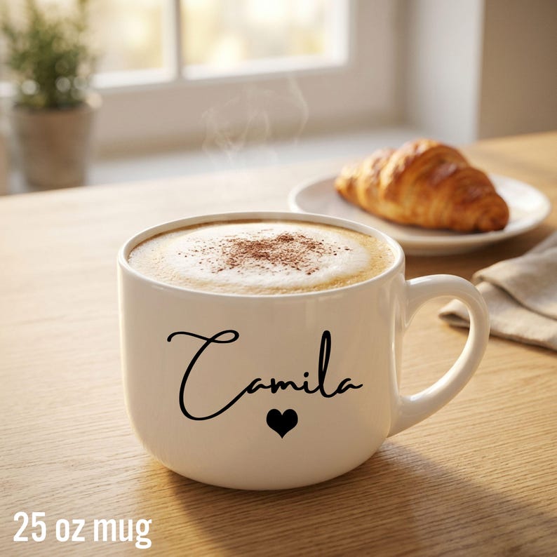 May include: A white 25 oz ceramic mug featuring the name Camila in black cursive script with a small black heart icon below the text. The mug is filled with a steaming hot beverage topped with a layer of foam and a light dusting of cocoa powder.