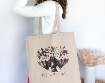May include: A beige tote bag with a black bat graphic and pink flowers. The text "Bal shil crazy" is printed on the bag.