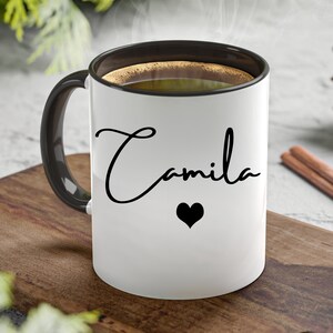 May include: A white ceramic mug with a black interior and a black handle. The exterior features the name Camila written in a black cursive script font, positioned above a small, solid black heart icon. The mug is filled with a dark brown liquid.