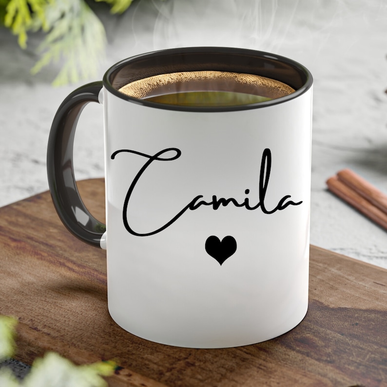 May include: A white ceramic mug with a black interior and a black handle. The exterior features the name Camila written in a black cursive script font, positioned above a small, solid black heart icon. The mug is filled with a dark brown liquid.
