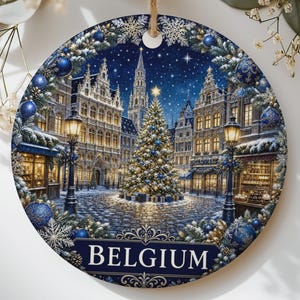 May include: A round ceramic ornament featuring a detailed illustration of a snow-covered town square in Belgium at night, centered by a glowing Christmas tree, framed by blue ornaments and snowflakes, with the word BELGIUM printed at the bottom.
