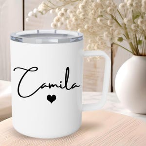 May include: A white cylindrical mug with a handle and a clear lid. The front of the mug features the name Camila written in a black cursive script font, positioned directly above a small, solid black heart icon centered on the smooth surface.