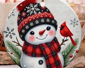 May include: A round Christmas ornament featuring a snowman with a red and black plaid scarf and hat. A red cardinal bird is perched on a branch next to the snowman. The ornament has a gold string for hanging.