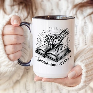 Puede incluir: Taza de cerámica blanca con borde y asa negros. La taza presenta una ilustración en blanco y negro de una mano esquelética sosteniendo un libro abierto. El texto "Spread those Pages" está impreso debajo del libro.