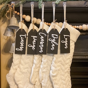 May include: Five white knitted stockings with black name tags hanging from a fireplace mantel. The name tags read "Timmy", "Kaige", "Cameron", "Ayden", and "Logan".