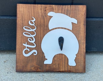 May include: A wooden sign with a white silhouette of a dog's rear end and a black hook. The name "Stella" is written in white cursive on the left side of the sign.