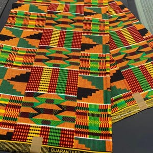 May include: A vibrant Kente cloth textile with a repeating geometric pattern in red, yellow, green, and black. The fabric is folded and laid out, showcasing the intricate design. The text "GUARANTEED EXCLUSIVE" is visible on the edge.