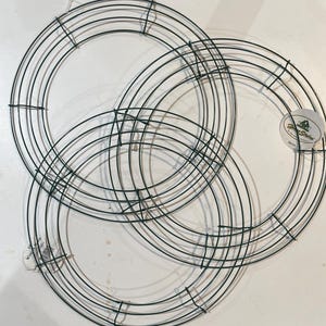 May include: Three green metal wreath frames overlapping each other. The frames are made of thin wire and are circular in shape. A white sticker with green text is visible on one of the frames. The frames are for crafting.