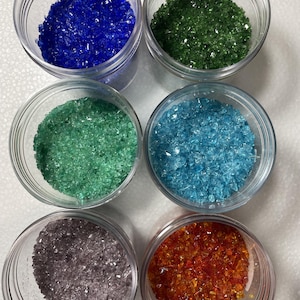 May include: Six clear plastic jars filled with small, angular crushed glass fragments in distinct colors: deep blue, forest green, light mint green, bright turquoise, muted lavender, and vibrant orange, arranged in a two-by-three grid pattern.