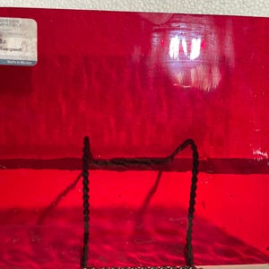May include: A sheet of vibrant red glass, labeled "Starry Red Waterglass" by Oceanside Compatible. The glass is supported by a black metal stand. The glass has a glossy finish and is made in Mexico.