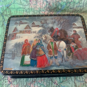 Puede incluir: Una caja de madera pintada a mano con una escena de invierno que representa un carruaje tirado por caballos y personas con ropa tradicional rusa. La caja está decorada con un borde negro y dorado.