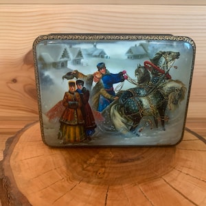 Pode incluir: Uma caixa de madeira pintada à mão com uma cena de inverno que retrata uma carruagem puxada por cavalos com três pessoas em roupas tradicionais russas. A cena se passa em um cenário de casas e árvores cobertas de neve.