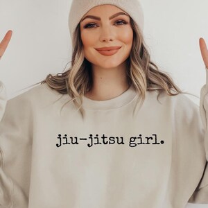 May include: A beige sweatshirt with the text "jiu-jitsu girl." printed in black on the front. The model is wearing a beige beanie and making a peace sign with both hands.