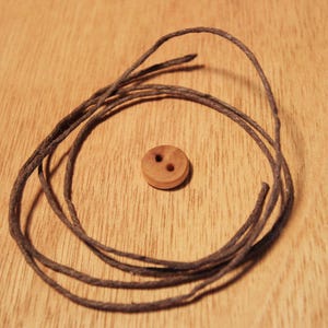 May include: A small, round, wooden button with two holes sits on a wooden surface. Dark brown twine is arranged in a loose circle around the button. The button and twine are the focal point of the image.