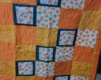 May include: A quilt with a patchwork design featuring orange, yellow, and white fabric. The fabric has a pattern of pumpkins and the words "Pumpkins" are printed on some of the fabric squares. The quilt is quilted with a swirl pattern.