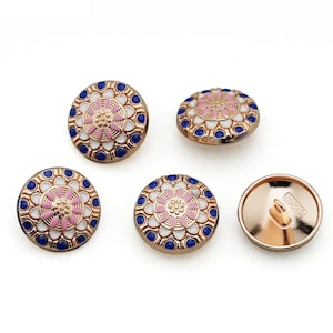 May include: A collection of five decorative buttons. The buttons are round with a gold-colored metal base. Each button features a central pink floral design, surrounded by white and blue accents. One button shows the back with a loop.