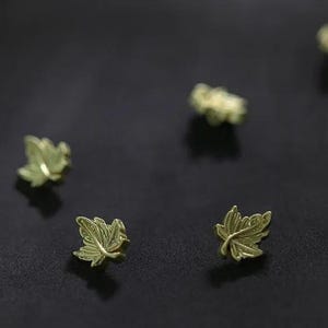 May include: Close-up of several small, gold-coloured leaf-shaped objects. The leaves have detailed veining and are scattered on a dark surface. The leaves appear to be made of metal.