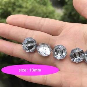 Op de afbeelding: Vijf heldere, gefacetteerde glazen knopen in zilverkleur. De knopen zijn 13 mm groot. De knopen zijn in een rij op een hand gerangschikt.