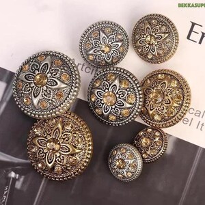 Puede incluir: Surtido de seis botones decorativos con acabado dorado y plateado. Cada botón presenta un diseño ornamentado en relieve con una estrella central y diamantes de imitación.