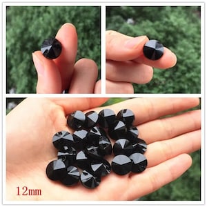 Puede incluir: Primer plano de cuentas de cristal negro de 12 mm con diseño facetado. Las cuentas están dispuestas en una pila sobre una mano.