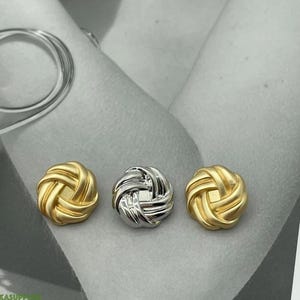 May include: Three decorative knot-shaped buttons. Two are gold-colored, and one is silver. The buttons have a woven design and are set against a neutral background. These buttons could be used for clothing or craft projects.