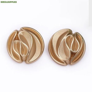Puede incluir: Dos botones de metal de color dorado con un diseño decorativo abstracto. Los botones tienen una superficie texturizada y ondulada.