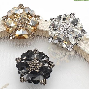 Può includere: Tre decorazioni in strass a forma di fiore in oro, argento e nero. Ogni decorazione ha un gruppo centrale di strass più piccoli.