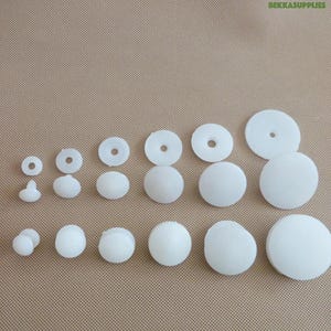 May include: An assortment of white plastic craft supplies, including round discs, spheres, and button-like shapes. The items vary in size and are arranged on a tan surface. The text "BEKKASUPPLIES" is visible in the top right corner.