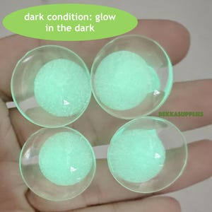 Op de afbeelding: Vier ronde, heldere glazen cabochons gevuld met een lichtgroen, in het donker lichtgevend poeder. De cabochons zijn in een vierkant op een hand gerangschikt. De tekst "dark condition: glow in the dark" is in groen op een witte achtergrond boven de cabochons geschreven.