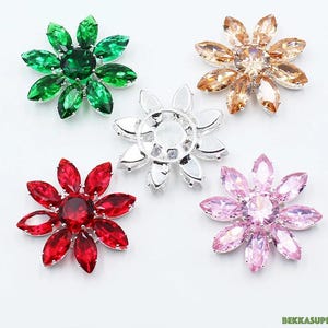 Puede incluir: Cinco adornos de strass en forma de flor con engastes plateados. Los strass son en tonos verde, marrón, transparente, rosa y rojo.