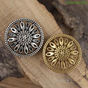 Może przedstawiać: Dwa ozdobne metalowe guziki z kwiatowymi wzorami. Jeden guzik jest srebrny, a drugi złoty. Guziki mają szczegółowy, wytłoczony wzór z centralnym kwiatem i elementami w kształcie płatków na obrzeżach. Idealne do projektów rzemieślniczych.