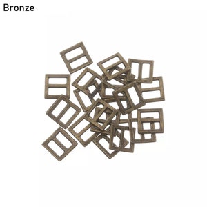 Op de afbeelding: Een stapel van 20 bronzen metalen schuifgespen. De gespen zijn vierkant met twee rechthoekige gaten in het midden.