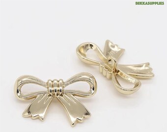 May include: Two gold-colored bow-shaped embellishments. The bows have a shiny finish and are detailed with a textured center and pointed ends. The background is plain white.