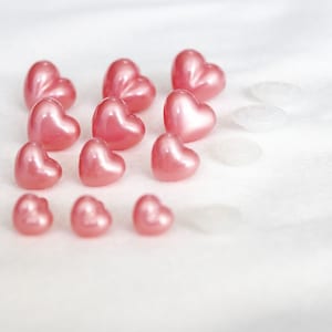 May include: A collection of pink heart-shaped buttons, some with a glossy finish, arranged on a white surface.