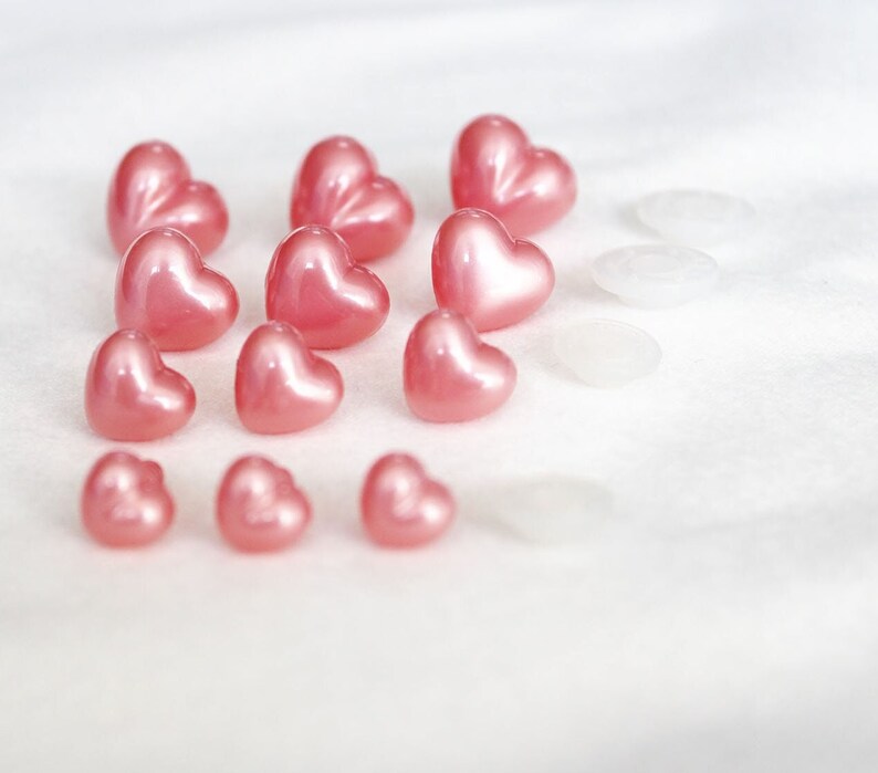 May include: A collection of pink heart-shaped buttons, some with a glossy finish, arranged on a white surface.