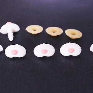 May include: A collection of plastic safety noses for stuffed animals, featuring white heart-shaped pieces with small pink centers, some with threaded stems and others with flat backs, alongside three tan circular washers for secure attachment.
