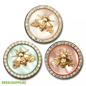 Puede incluir: Tres botones redondos decorativos con un diseño de abeja dorada. Cada botón tiene un fondo de color diferente: blanco, verde menta y rosa. Los botones están rodeados por un anillo de pequeños cristales transparentes y un borde de metal dorado.