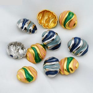 Può includere: Una collezione di perline decorative in vari design e colori. Alcune perline sono a forma di conchiglia, mentre altre sono rotonde con strisce smaltate oro, rosa, verdi e blu. Le perline sono disposte su una superficie bianca.